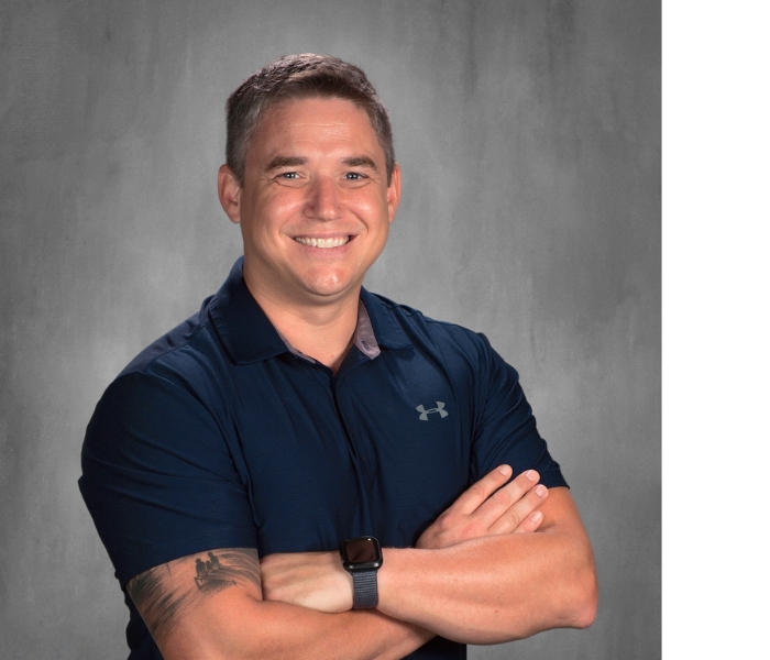 A man with short, graying hair and a beard smiles with his arms crossed. He is wearing a dark blue polo shirt with a gray collar and an Under Armour logo on the chest. He has a tattoo on his left arm and is wearing a smartwatch.