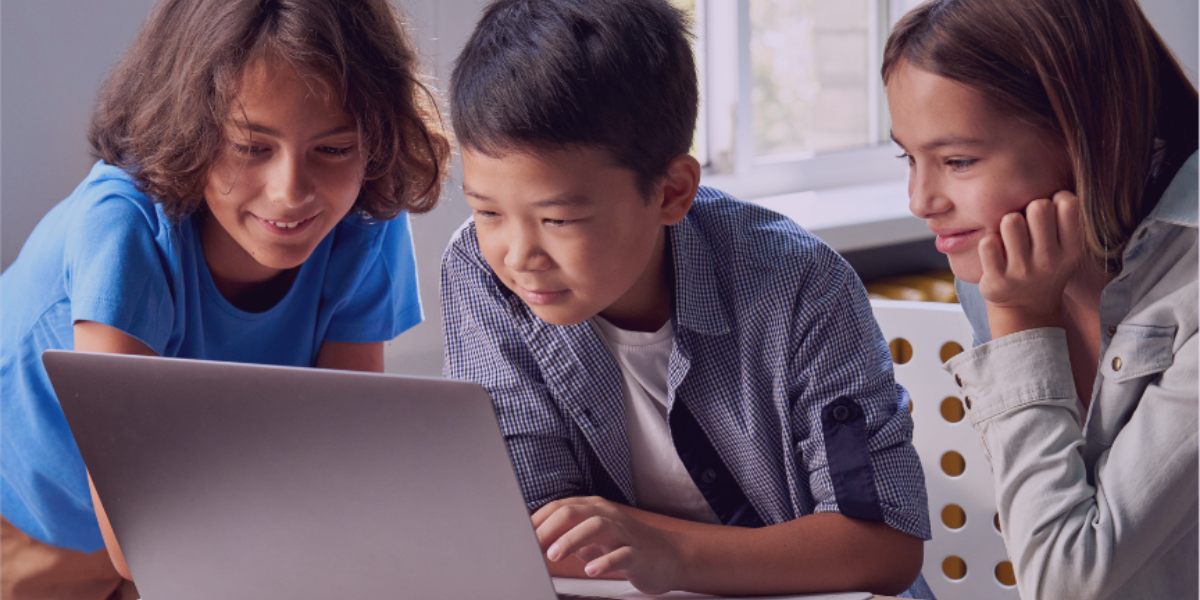 The image shows three children looking at a laptop computer with an intrigued, concentrated expression.