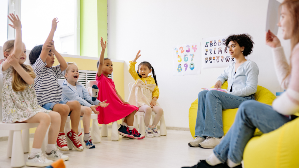 Children seated in chairs, eagerly raising their hands in a classroom setting.