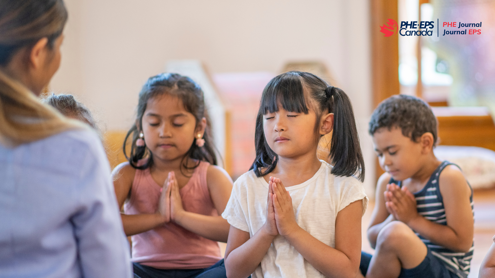 The image shows three children practicing mindfulness with praying hands, eyes closed, in front of their teachers.