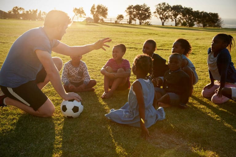 physical education teacher holding a soccer ball talking to young children while sitting on a grass field