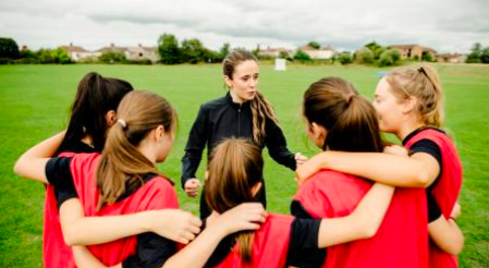 group of 5 young women in a huddle wearing red jerseys and a coach in the centre wearing a black jacket on a soccer field