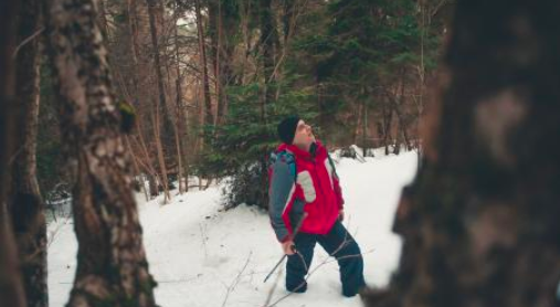 man standing on snow in the woods looking up wearing a red winter jacket. 