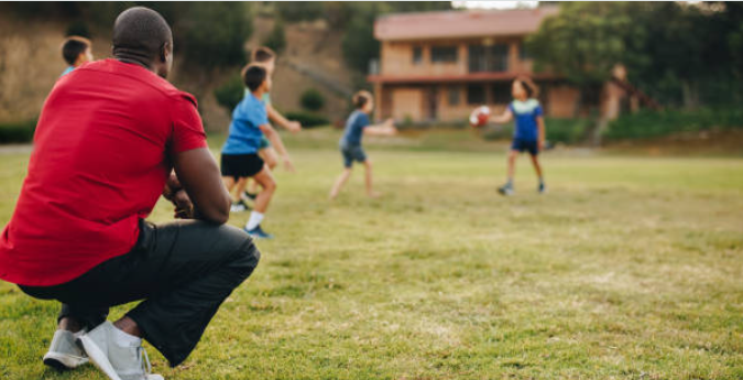 a physical education teacher squating in a grass field watching their students run an activity