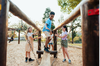 young Black child walking across a balance beam on a play structure outside with friends on either side supporting him