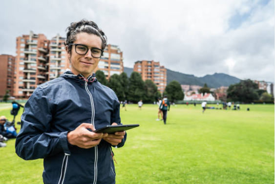 a man holding an ipad wearing glass and a black windbreaker standing on a grass field smiling. Looks like a coach holding an ipad to keep track of his players. 