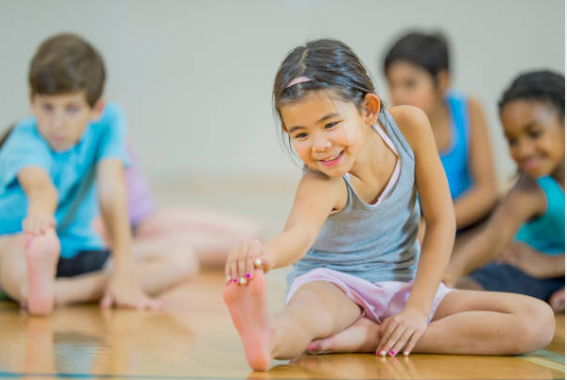 a young child in a gymnasium sitting on the ground stretching their leg. There are other children in the background doing the same stretch trying to touch their toes. 