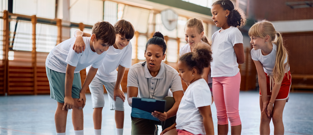 there is a light skinned, woman, physical education teacher in the centre down on one knee. She is holding a clipboard, and has two boys students to her right bent over looking at the clip board. On the teacher's left, she has 4 young girls looking at the clipboard. 3 of them and bent over looking down and there is one on her knees. 