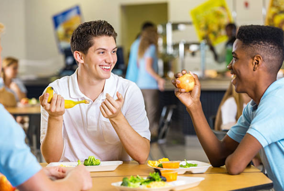 two teenage boys sitting at a cafeteria table. One is holding a banana and smiling at the other boy who is smiling and holding an apple. There are plates on the table that have broccoli and other green vegetables. 
