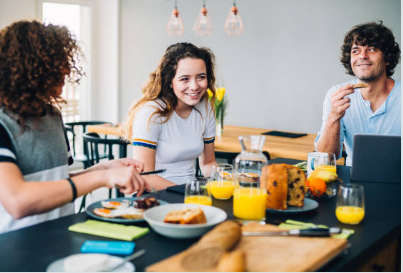3 teenagers sitting at a table eating breakfast. The table has many breakfast food items like eggs, toast, and orange juice. 