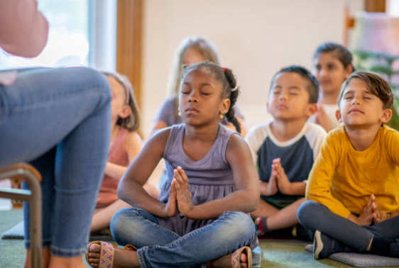 student sitting cross legged on the floor in their classroom. Their palms are touching and fingers pointed to the sky as their arms rest on their knees. They are meditating. 