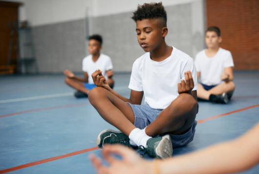 3 students sitting cross legged on a gym floor. They have their index finger and thumb touching while their hand rests on their knees. Their eyes are closed and they are meditating. 