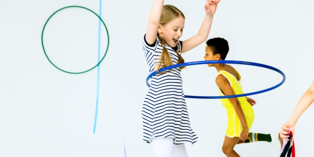 Three children are playing with hula hoops and ribbons against a white background. One child in a striped dress is hula hooping, another child in a yellow outfit is running, and a third child's arm is visible holding ribbons.