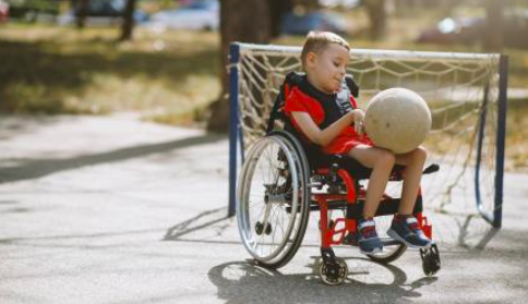boy in wheelchair wearing a red shirt and red shorts holding a soccer ball with a net behind him