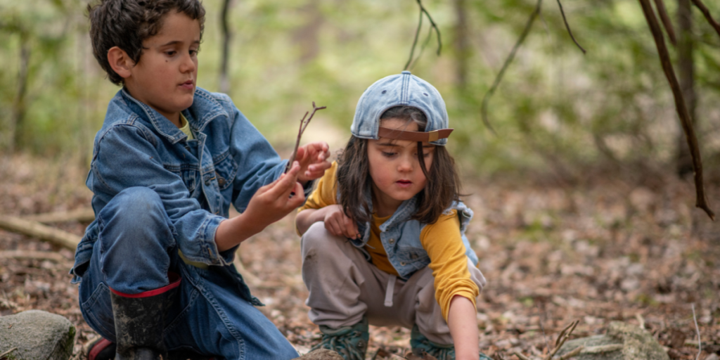 Two children crouching in a forest, examining twigs and rocks on the ground.