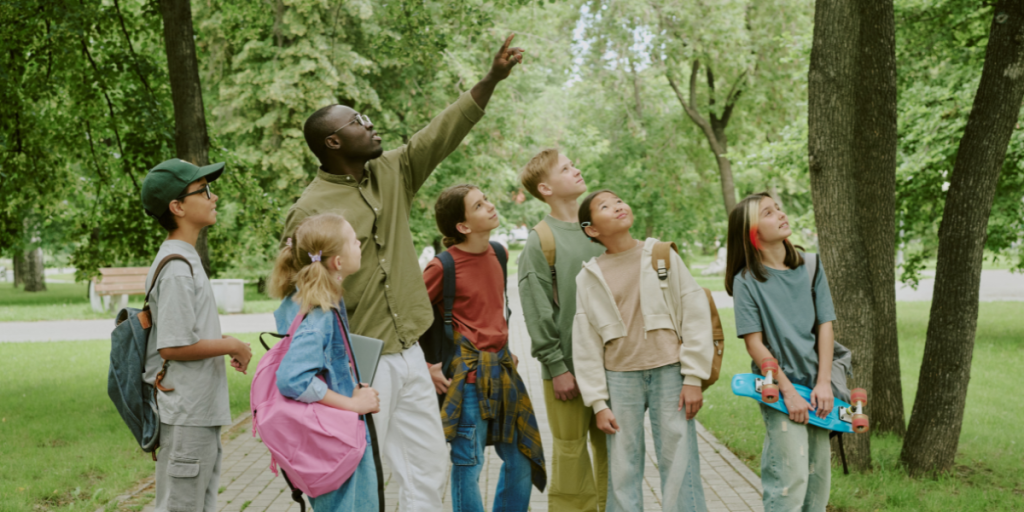 A man points upwards while a group of children look in the same direction in a park setting.