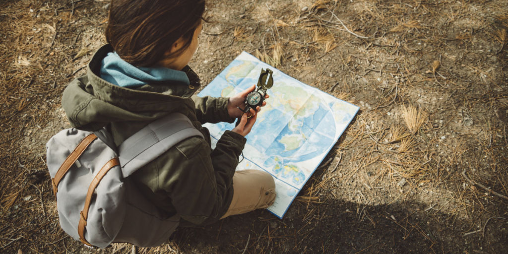 A person wearing a green jacket and backpack holds a compass over a map spread on the ground.