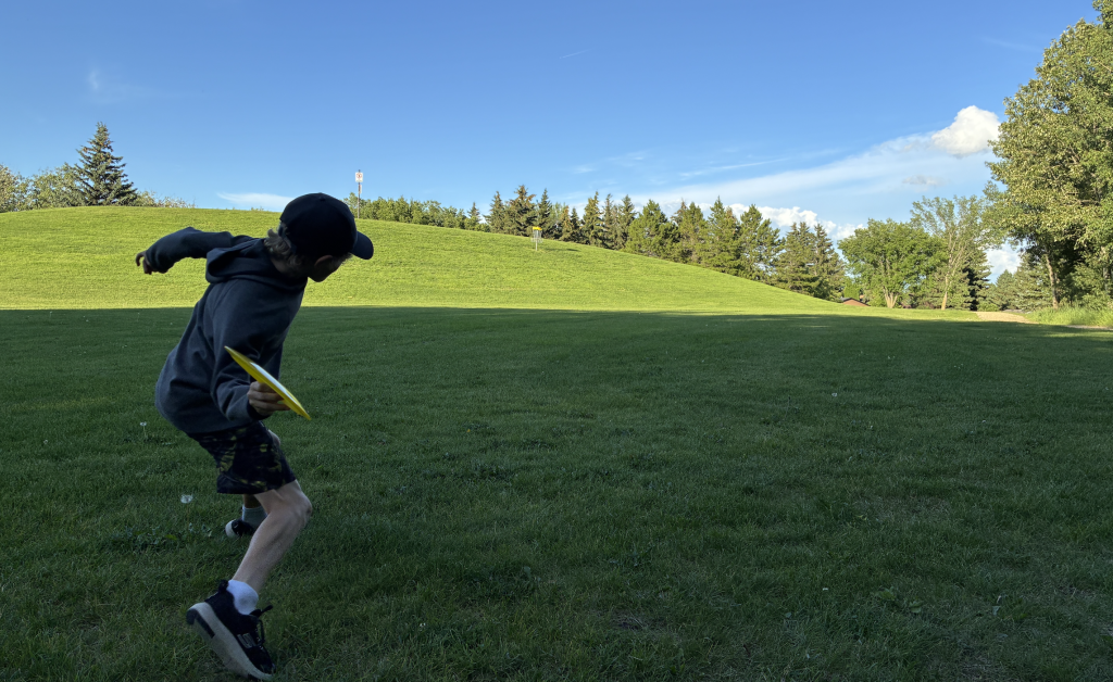 a kid is doing disc golf in a outdoor green space under bright blue sky
