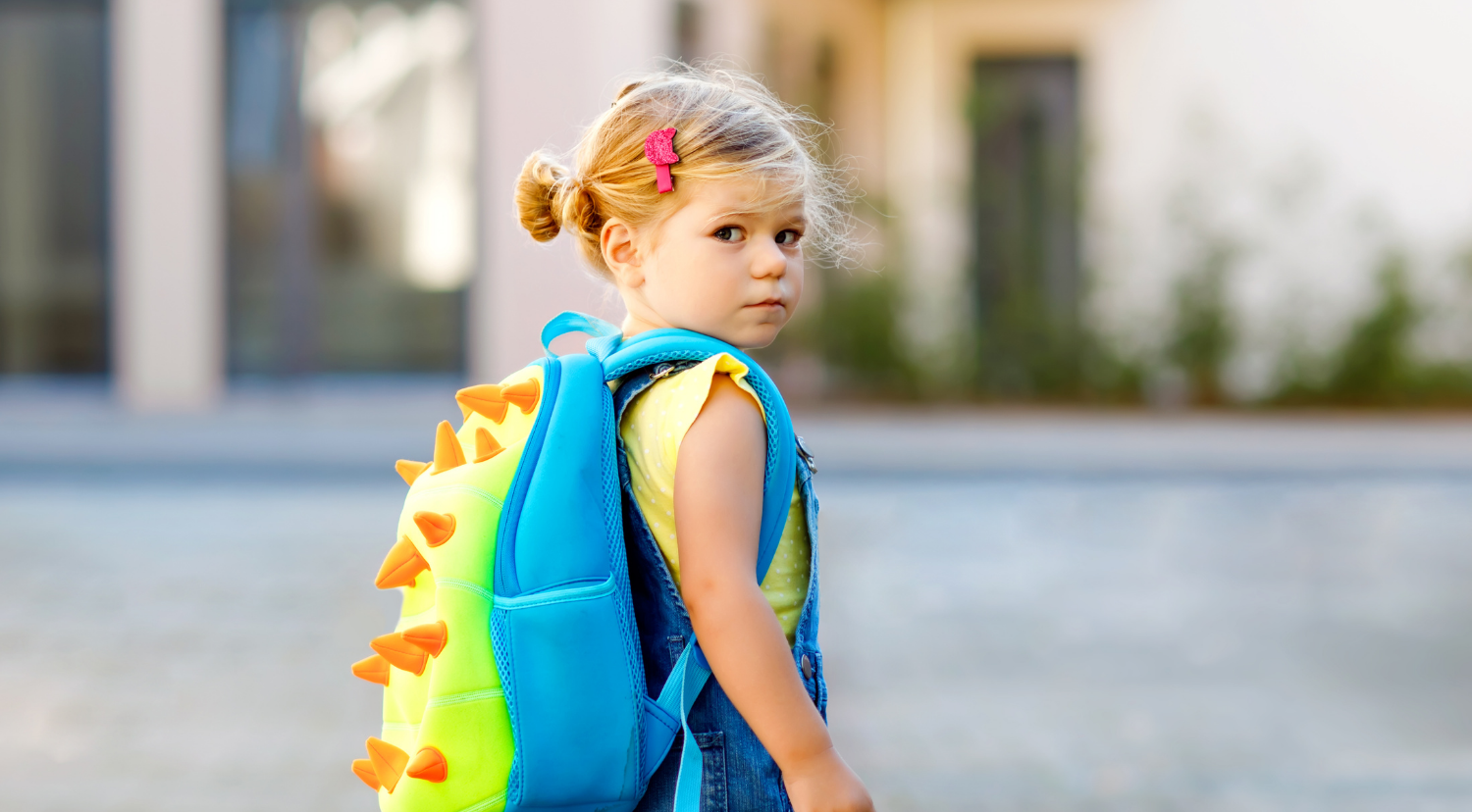 A young child with blonde hair in a bun, wearing denim overalls and a yellow shirt, looks over their shoulder while carrying a bright blue and green dinosaur backpack with orange spikes.