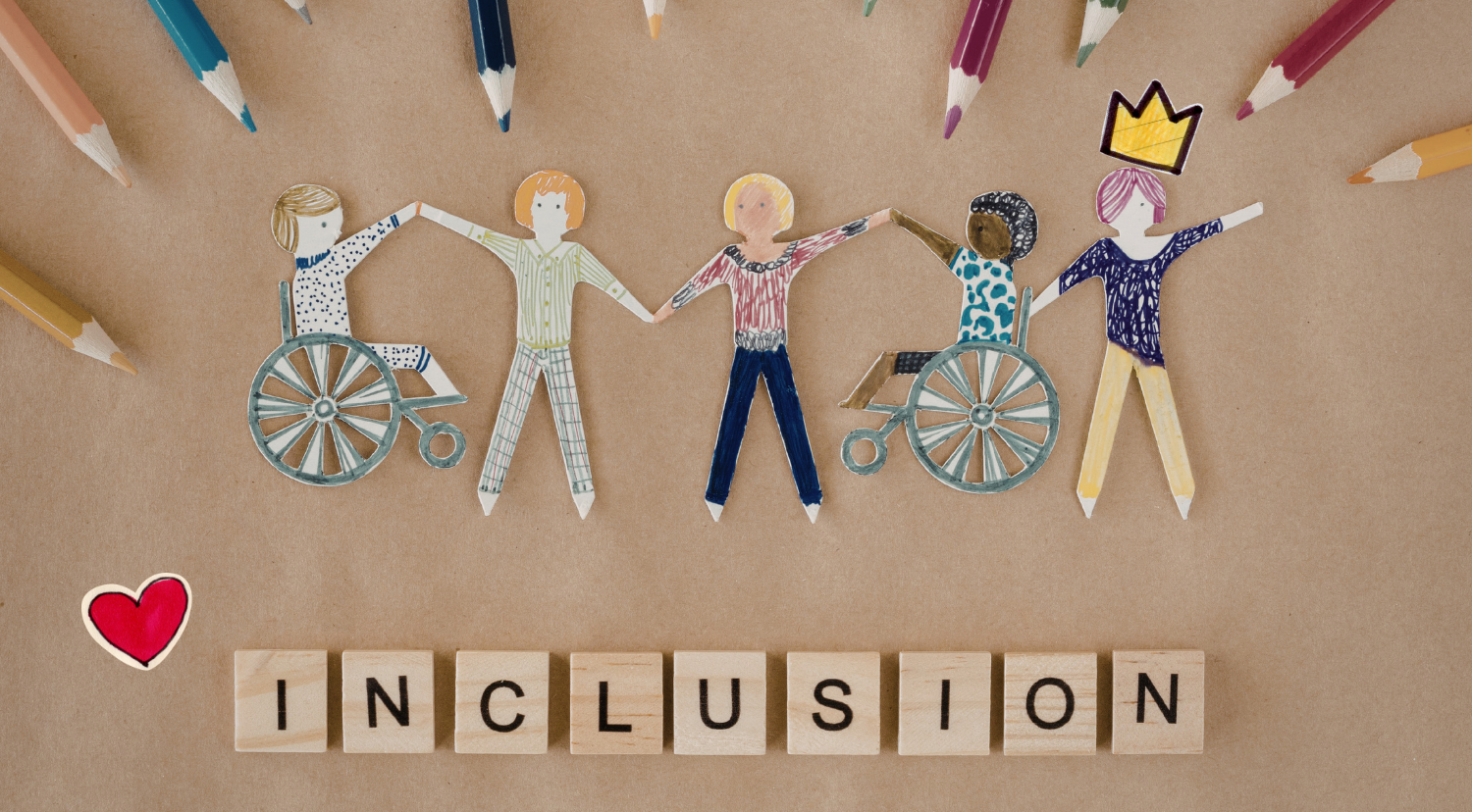 Paper cutouts of diverse people, including some in wheelchairs, holding hands, surrounded by colored pencils. Wooden letter blocks spell out 'INCLUSION'.