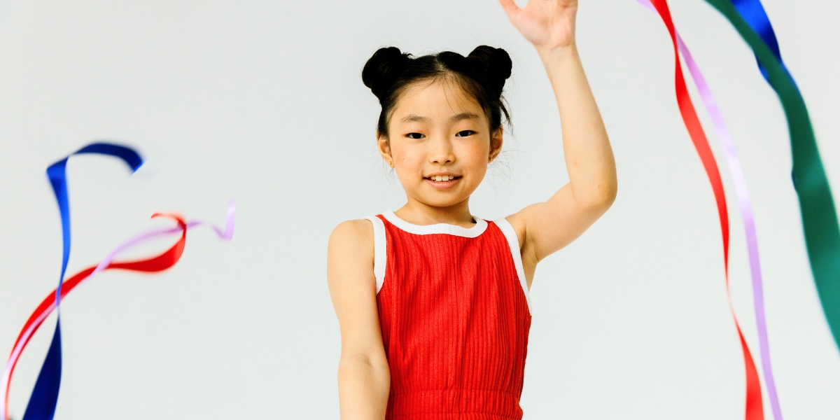 A young person in a red leotard smiles while holding up a hand and waving colorful ribbons against a white background.