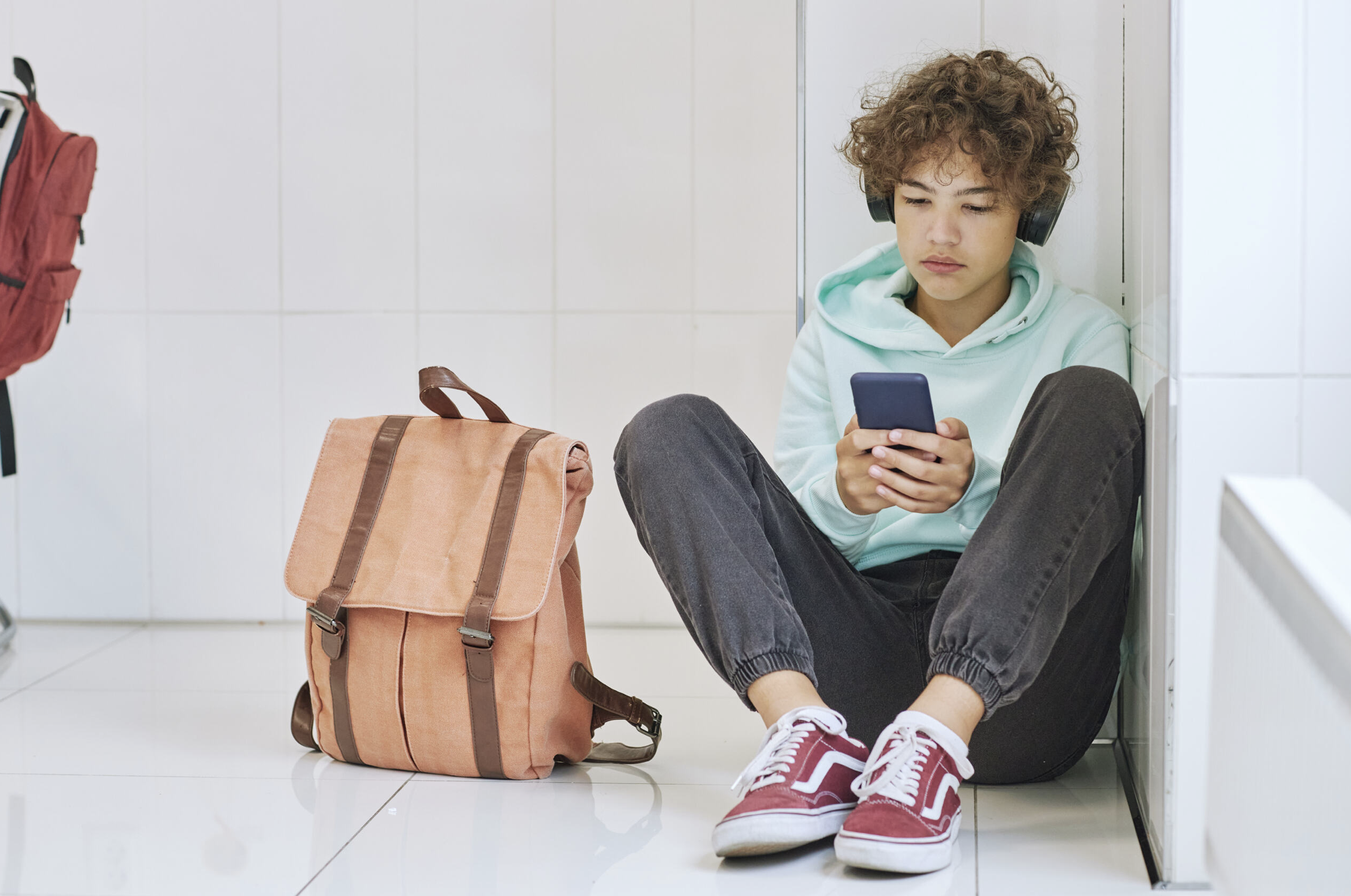 A teenage boy is sitting on the floor with a schoolbag next to him. He is holding a phone and looking at it intently.