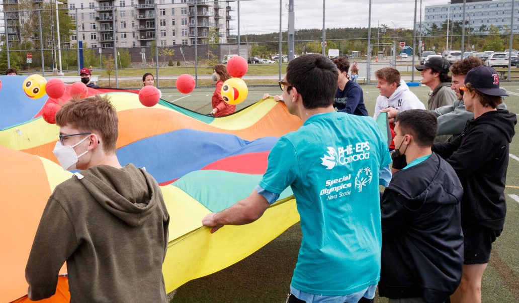 students in a field holding onto a colour parachute pulling it up and down for red and yellow dodgeballs to fly into the air.