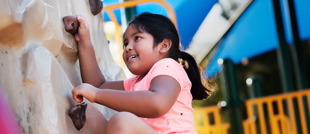 a young brown girl climbing on a rock climbing wall. She is wearing a pink shirt, and has medium length black hair in pigtails. 