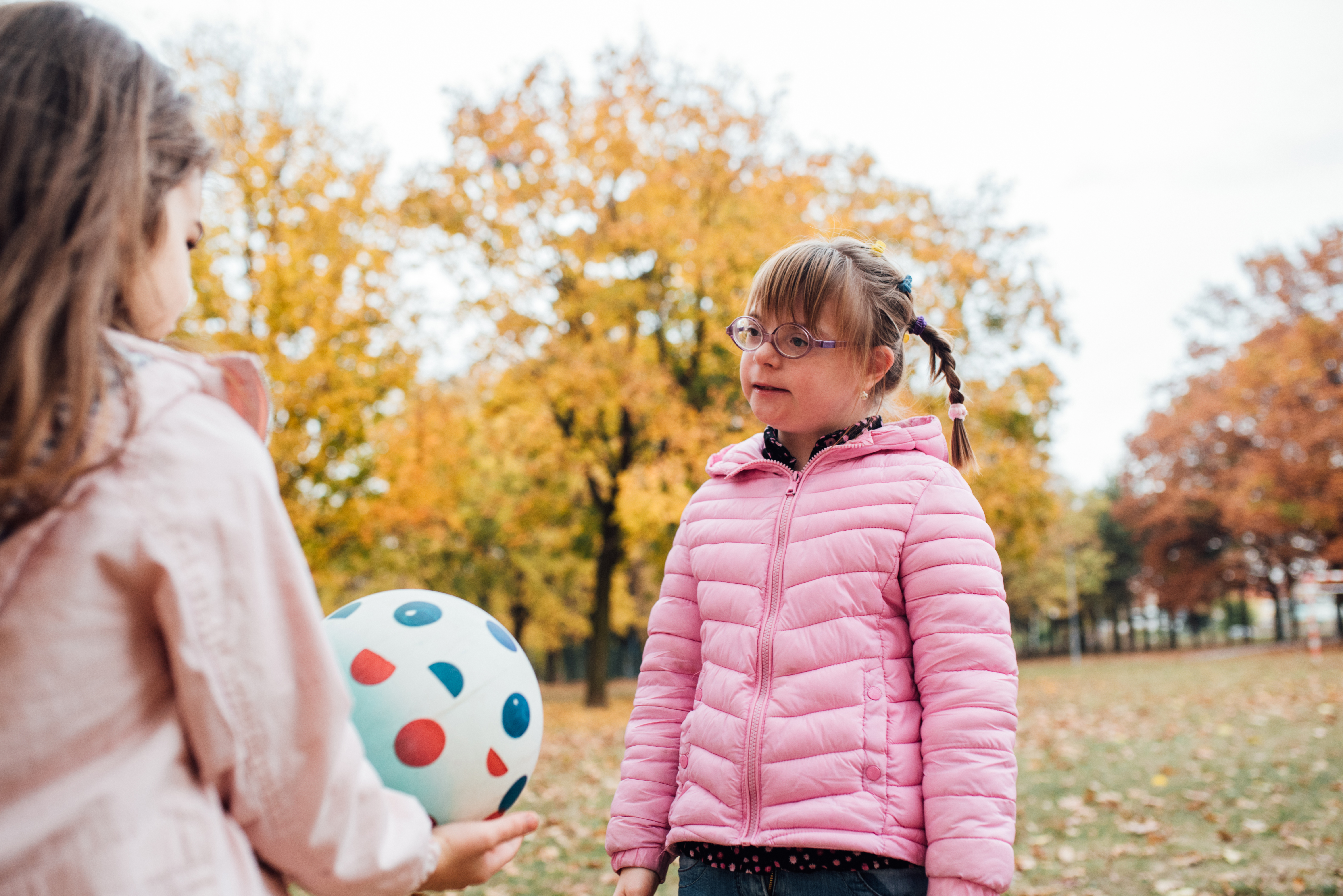 a child with down syndrome wearing a pink puff jacket smiling at an adult with blonde hair wearing a off white jacket holding a soccer ball