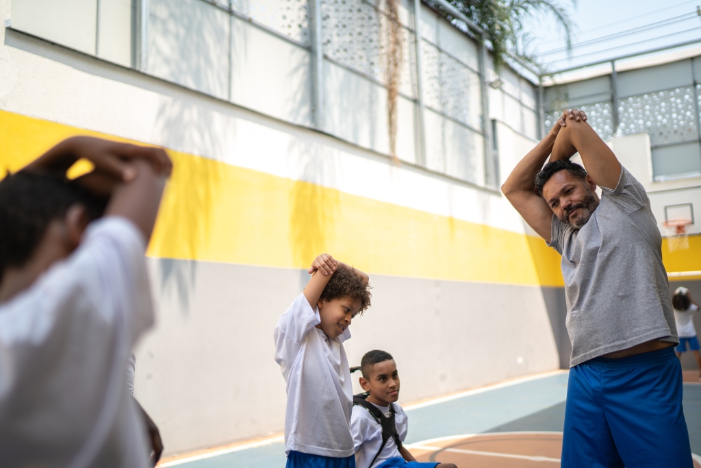 a black man who is a physical education teacher is stretching with 3 of his students. Two are standing with one arm over their head and another is in a wheelchair.