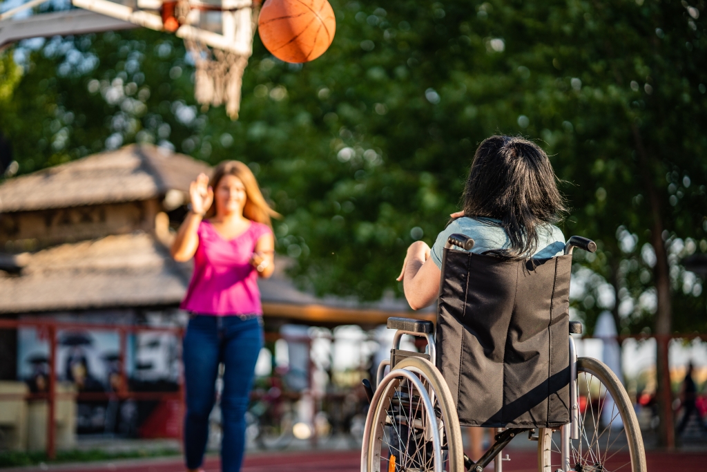 child in a wheelchair shooting a basketball at a basketball net and an adult standing under the net ready to catch the ball