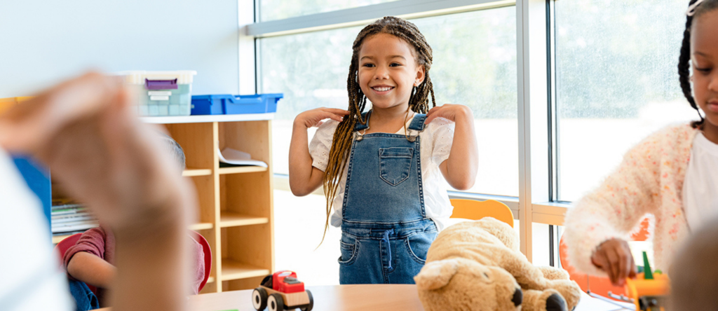 a young black girl standing behind a desk with her hands on her shoulder. She is smiling and there is a toy car on the desk and a teddy bear. 