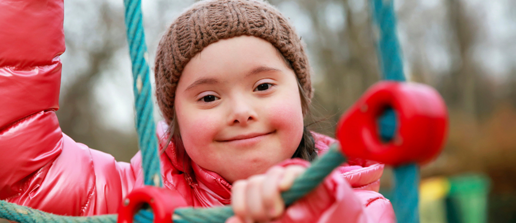 a young girl with down syndrome is smiling. She is at a playground and is wearing a brown toque, and pink puffer jacket. 