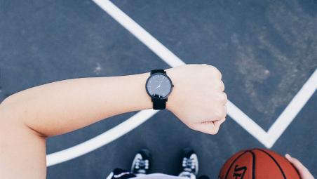 a person holding a basketball and looking at their watch. You can only see their forearm, hand, shoes and half the basketball