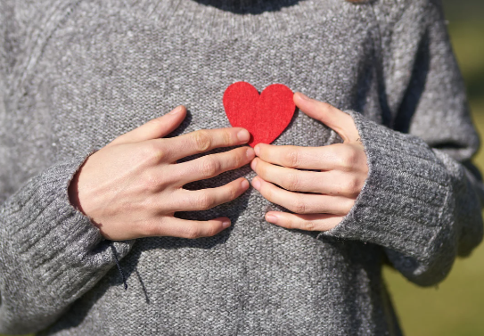 A person in a grey sweater holding a red paper-cut heart, symbolizing self-love.
