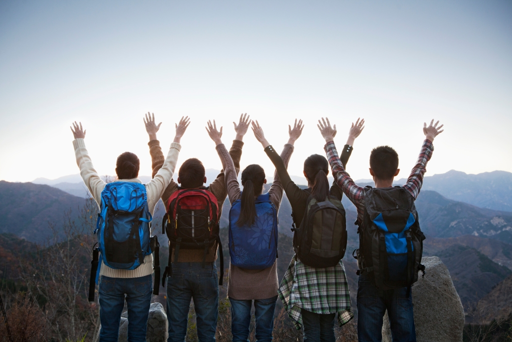 5 students on the edge of a cliff wearing backpacks, with their hands up 