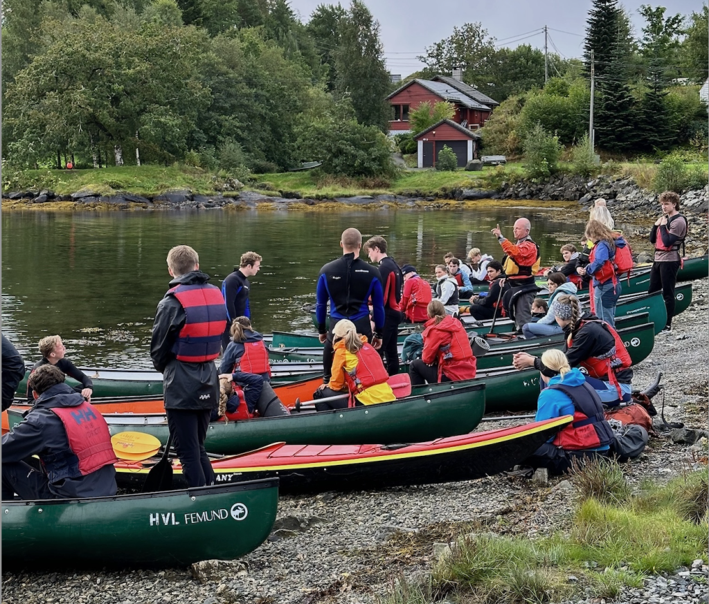 The photo shows a group of students preparing for an outdoor kayak/canoe course in a picturesque area.