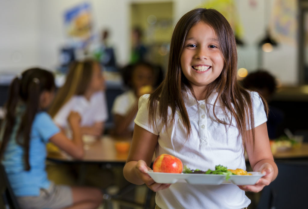A young girl holding a plate of food in what appears to be a school canteen 