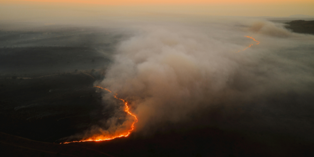 An aerial view of a wildfire at sunset, with flames and smoke rising from the ground and a warm orange glow in the sky.