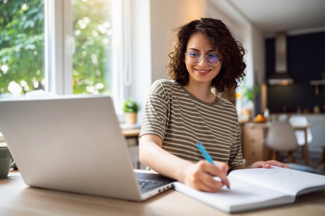 A woman wearing glasses sits at a table, working on a laptop with a notebook beside her.