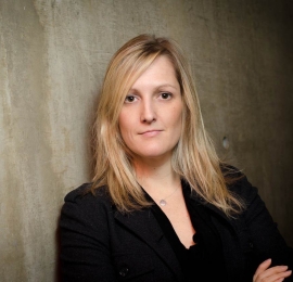 middle aged, white woman, with shoulder length blonde harid. She is wearing a black, long sleeved blouse, and a silver necklace. She is standing with her arms crossed against a cement wall.