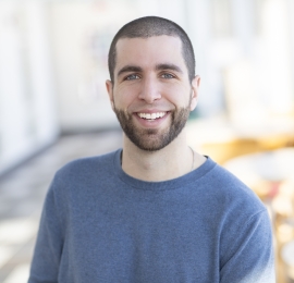 White male, around age 30, with brown buzz cut hair and a dark brown beard. He has light blue eyes, and is standing outside smiling and wearing a light blue crew neck sweater. 