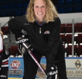 white woman with curling blonde midlength hair. She is wearing a black track suit and is standing on an ice rink holding a hockey stick while wearing hockey gloves. 