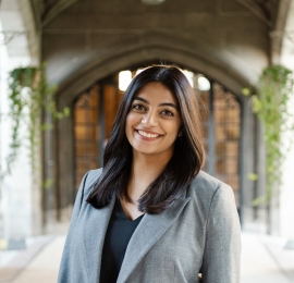 a woman, with brown skin tone, with shoulder-length dark brown hair. She is wearing a slate grey pea-coat, and a black v-neck undershirt. She is standing and smiling in front of a cement archway. 