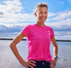 a middle-aged white woman, with blonde hair in a pony-tail, standing in a pink t-shirt with her hands on her hip infront of a lake