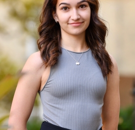 a woman with shoulder length brown hair with tanned skin tone. She is wearing a black adn white stripped tank top and dark blue pants. 