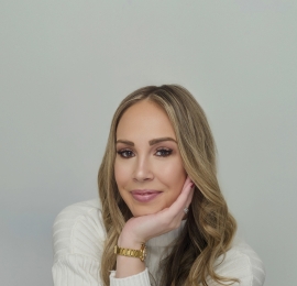 a white skin tone woman, with long blonde hair. She is wearing a white, long sleeved, cashmere shirt and a gold watch. She is sitting down, with her arms cross in fron of her with her chin resting in the pal, of her left hand.