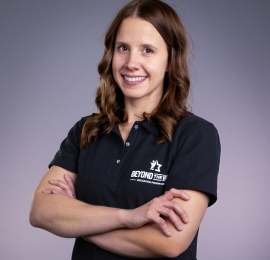 white women with shoulder length brown hair, smiling in the photo, she is standing with her arms crossed at her chest. She is wearing a black shirt and it has a small logo on the left chest that says "Beyond the Win - Education Program"