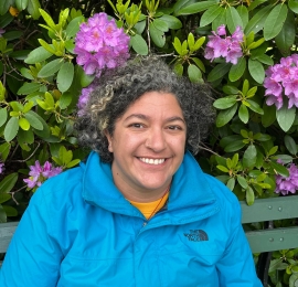 woman with dark and light grey curly hair, light brown skin, and is wearing a bright blue windbreaker with a yellow t-shirt underneath. There are pink/purple flowers and greenery in the back. 