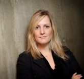 middle aged, white woman, with shoulder length blonde harid. She is wearing a black, long sleeved blouse, and a silver necklace. She is standing with her arms crossed against a cement wall.